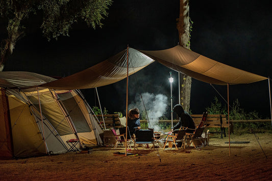 A nighttime camping scene with two people seated at a camp table under a tarp canopy, beside a large tent, illuminated by a standing camping lantern. Smoke rises in the air, suggesting a recently extinguished campfire or cooking.
